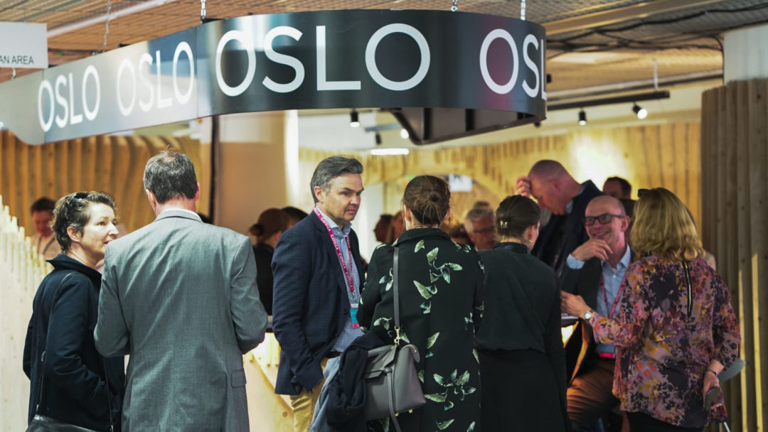 Mennesker samlet rundt en stand på et konferanseområde innendørs. Standen er merket med