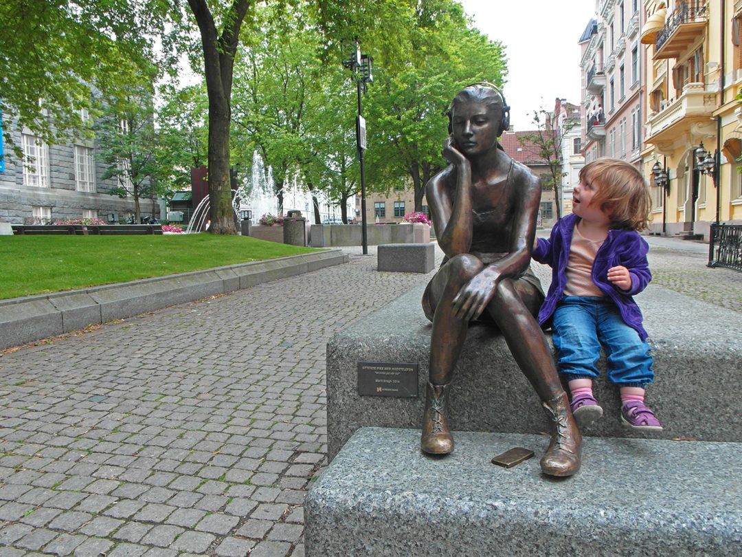 En liten jente sitter ved siden av en statue av en tenåringspike med hodetelefoner.